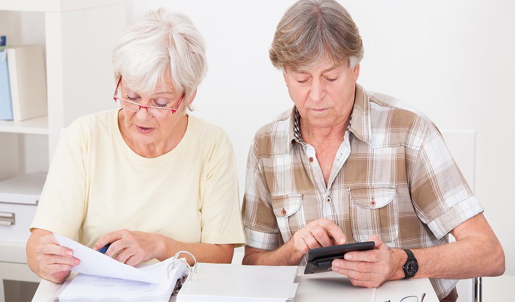Elderly Woman Checking The Accounts While The Husband Adds Up The Figures On The Calculator (Foto: Paulo Emílio) Elderly Woman Checking The Accounts While The Husband Adds Up The Figures On The Calculator