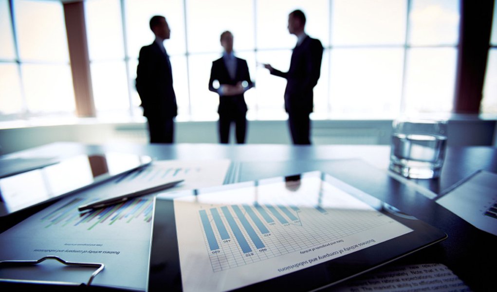 Close-shot of a tablet computer displaying financial data, three businessmen standing in the background (Foto: Paulo Emílio) Close-shot of a tablet computer displaying financial data, three businessmen standing in the background