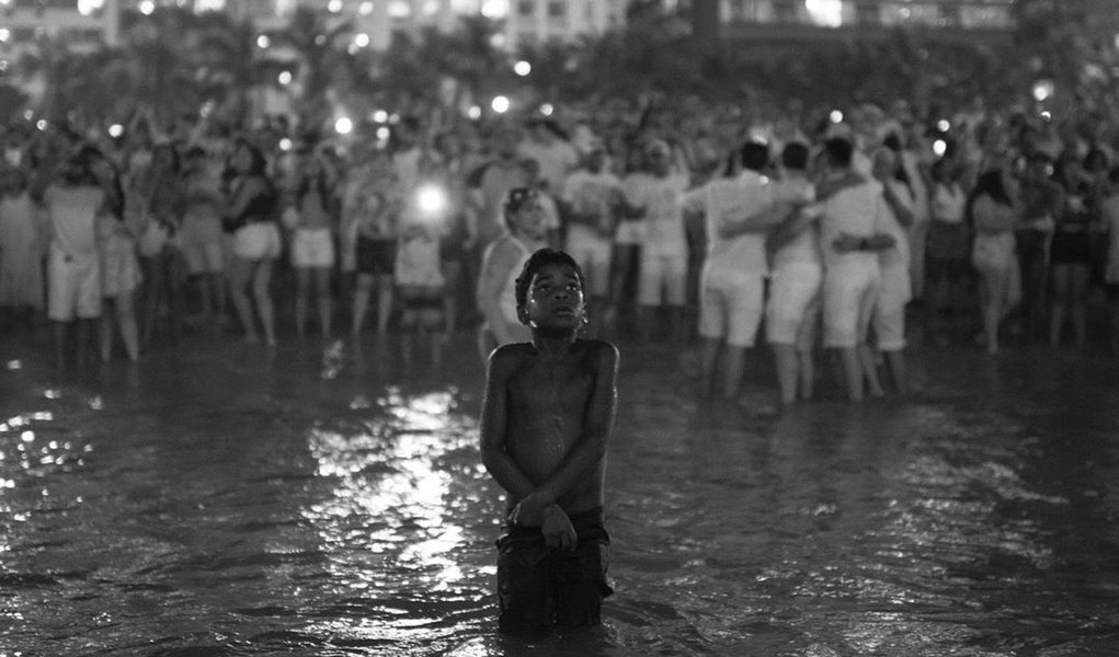 Imagem do fotógrafo Lucas Landau mostra um garoto negro e com frio dentro do mar, à frente de um grande grupo de pessoas vestidas de branco durante a queima de fogos do Ano Novo no Rio de Janeiro; postagem já teve mais de 20 mil reações e mais de 5 mil compartilhamentos; "A fotografia abre margem para várias interpretações; todas legítimas", comentou Landau, depois da repercussão (Foto: Gisele Federicce) Imagem do fotógrafo Lucas Landau mostra um garoto negro e com frio dentro do mar, à frente de um grande grupo de pessoas vestidas de branco durante a queima de fogos do Ano Novo no Rio de Janeiro; postagem já teve mais de 20 mil reações e mais de 5 mil compartilhamentos; "A fotografia abre margem para várias interpretações; todas legítimas", comentou Landau, depois da repercussão