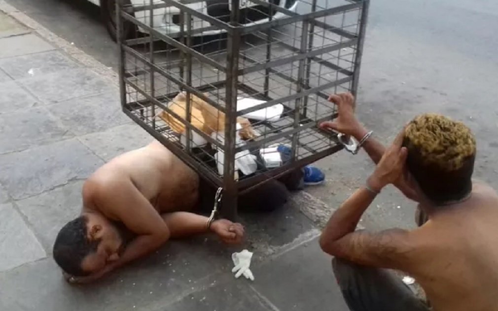 Uma cena inusitada chamou a atenção na manhã desta quarta-feira em Porta Alegre; dois presos que aguardavam vaga no sistema penitenciário foram algemados em uma lixeira, que fica em frente ao Palácio da Polícia; de acordo com os policiais que faziam a custódia dos presos, os dois pediram para esticar as pernas, uma vez que estão dentro da viatura desde a terça-feira; Rio Grande do Sul passa por uma crise no sistema carcerário, que faz com que os presos sejam mantidos dentro das viaturas por conta da falta de vagas (Foto: José Barbacena) Uma cena inusitada chamou a atenção na manhã desta quarta-feira em Porta Alegre; dois presos que aguardavam vaga no sistema penitenciário foram algemados em uma lixeira, que fica em frente ao Palácio da Polícia; de acordo com os policiais que faziam a custódia dos presos, os dois pediram para esticar as pernas, uma vez que estão dentro da viatura desde a terça-feira; Rio Grande do Sul passa por uma crise no sistema carcerário, que faz com que os presos sejam mantidos dentro das viaturas por conta da falta de vagas
