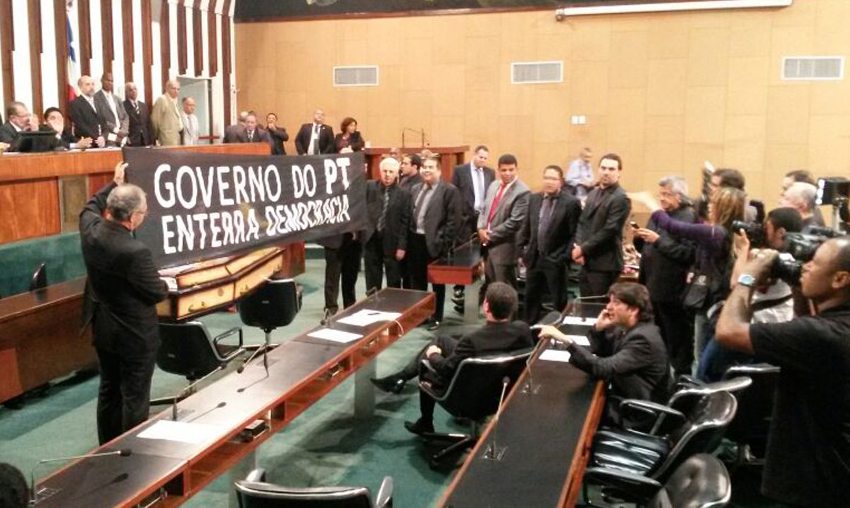 Protestando com um caixão e com 'velório' da democracia, que teria sido 'assassinada' pelo PT, os deputados da minoria na Assembleia Legislativa tentaram ontem anular a sessão que escolheu o deputado federal Zezéu Ribeiro, do PT, como novo conselheiro do Tribunal de Contas do Estado (TCE);o vice-líder da oposição, Bruno Reis (PMDB), apontou "diversas irregularidades no processo eleitoral", como as fotos feitas por governistas no momento do voto, "numa clara demonstração de que precisavam comprovar a escolha pelo candidato petista", em detrimento a Carlos Gaban (DEM) (Foto: Romulo Faro) Protestando com um caixão e com 'velório' da democracia, que teria sido 'assassinada' pelo PT, os deputados da minoria na Assembleia Legislativa tentaram ontem anular a sessão que escolheu o deputado federal Zezéu Ribeiro, do PT, como novo conselheiro do Tribunal de Contas do Estado (TCE);o vice-líder da oposição, Bruno Reis (PMDB), apontou "diversas irregularidades no processo eleitoral", como as fotos feitas por governistas no momento do voto, "numa clara demonstração de que precisavam comprovar a escolha pelo candidato petista", em detrimento a Carlos Gaban (DEM)