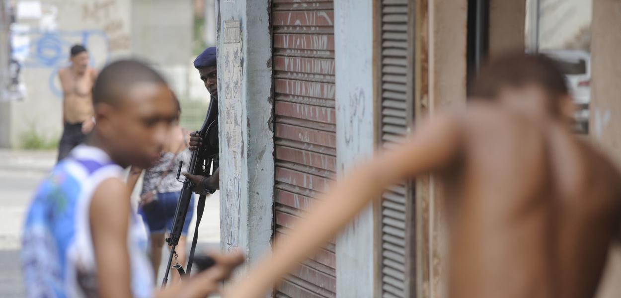 Policial armado observa jovens negros na favela (Foto: Tânia Rêgo/ Agência Brasil) Policial armado observa jovens negros na favela