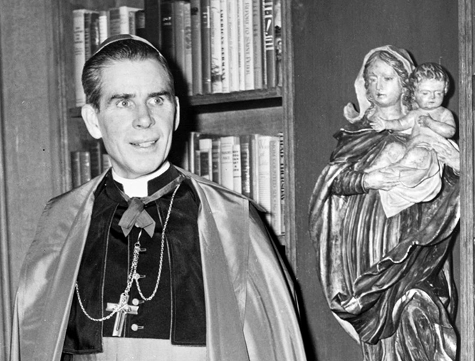 Bishop Fulton Sheen stands before a bookcase on the set of his Dumont television program. Bishop Fulton Sheen stands before a bookcase on the set of his Dumont television program.