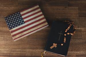 A table holds an American flag next to a Bible with a Rosary resting on it. A table holds an American flag next to a Bible with a Rosary resting on it.
