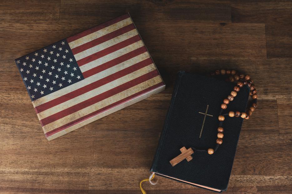 A table holds an American flag next to a Bible with a Rosary resting on it. A table holds an American flag next to a Bible with a Rosary resting on it.