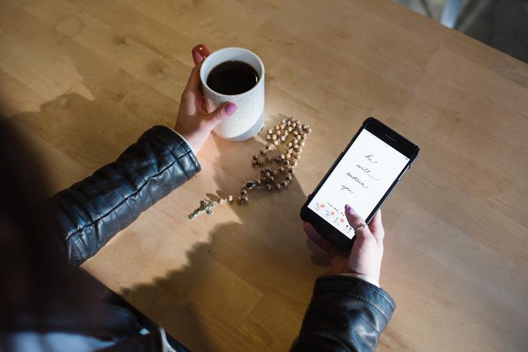 A woman peruses her phone while her Rosary sits on the table. A woman peruses her phone while her Rosary sits on the table.